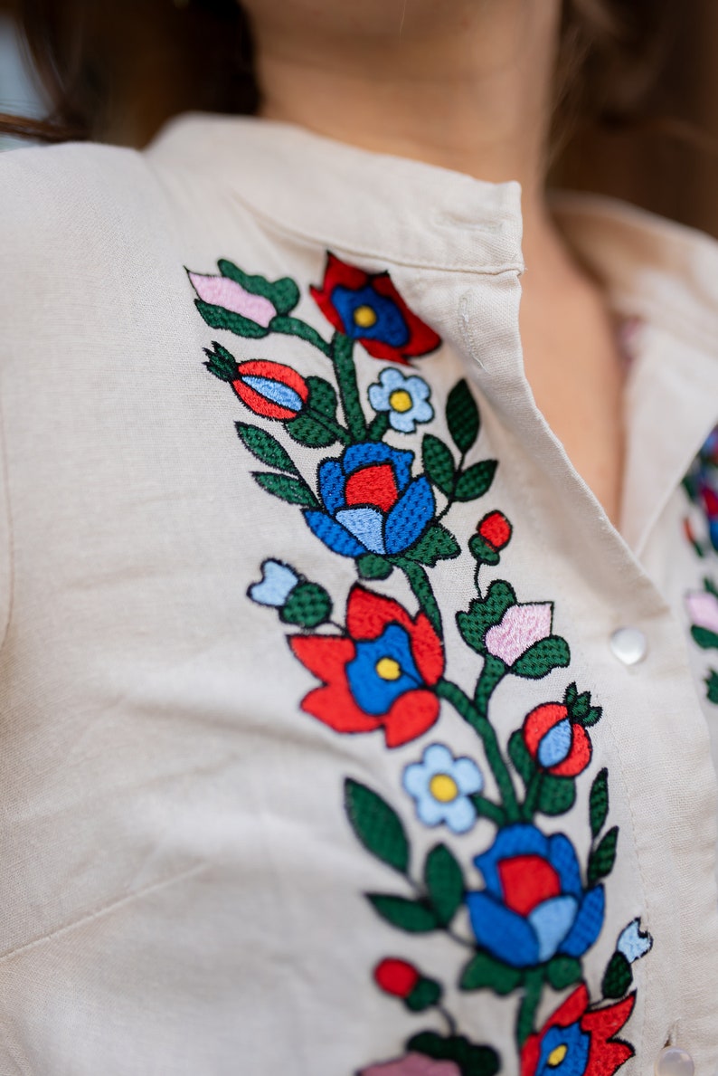 May include: Cream-colored shirt with colorful floral embroidery. The embroidery features red, blue, and pink flowers with green leaves. The shirt has a button-down front and a collar.