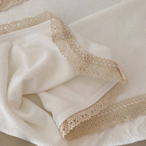May include: A white towel with a beige lace trim. The towel is folded in half and the lace trim is visible on both sides.