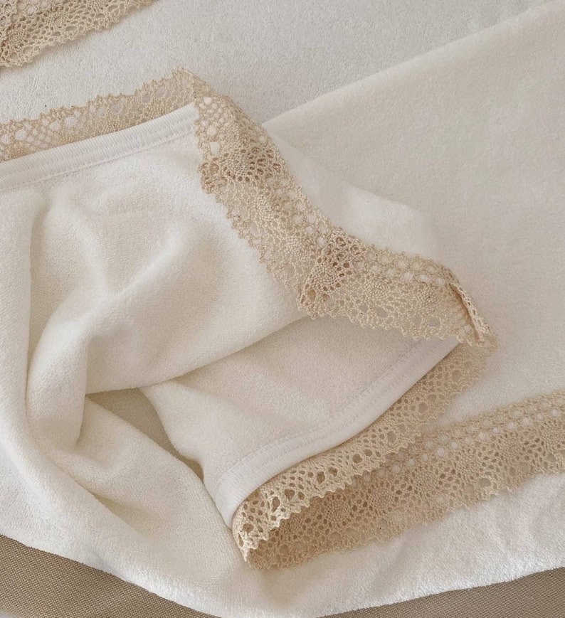 May include: A white towel with a beige lace trim. The towel is folded in half and the lace trim is visible on both sides.