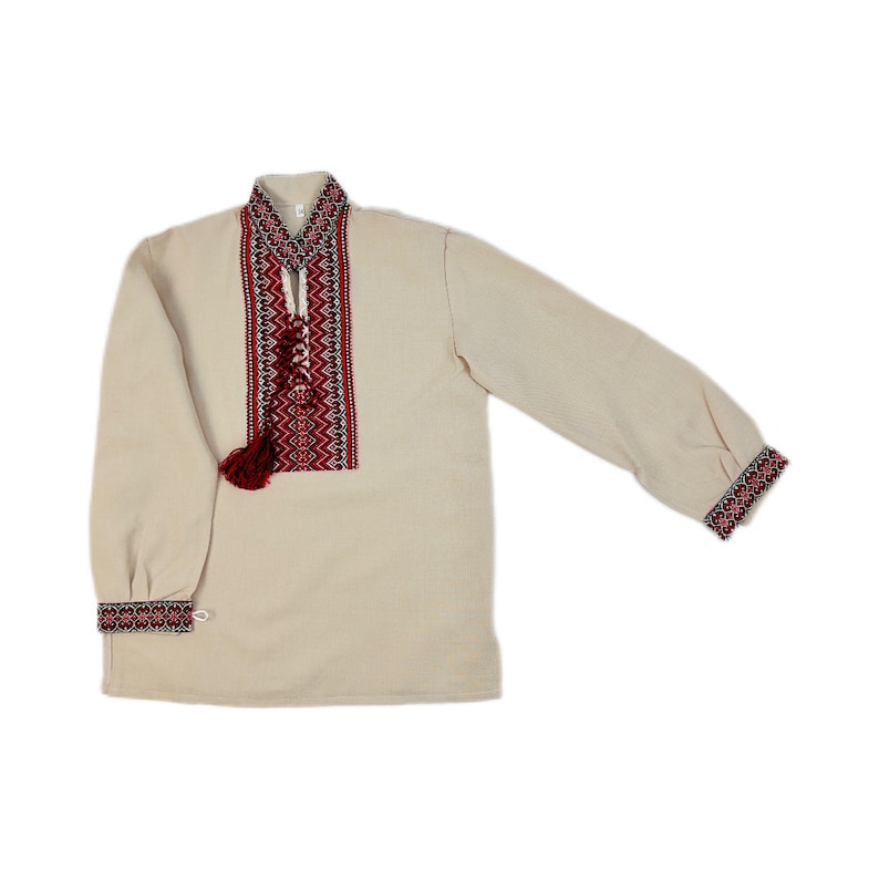 Op de afbeelding: Een cr&egrave;mekleurig shirt met lange mouwen met ingewikkelde rode en zwarte borduursels rond de kraag en over de voorkant. De manchetten hebben ook bijpassende borduursels. Een kwast hangt van de halslijn.