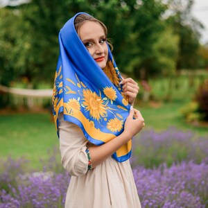 May include: A woman wearing a blue and yellow patterned scarf with sunflowers. She is standing in a field of lavender flowers.