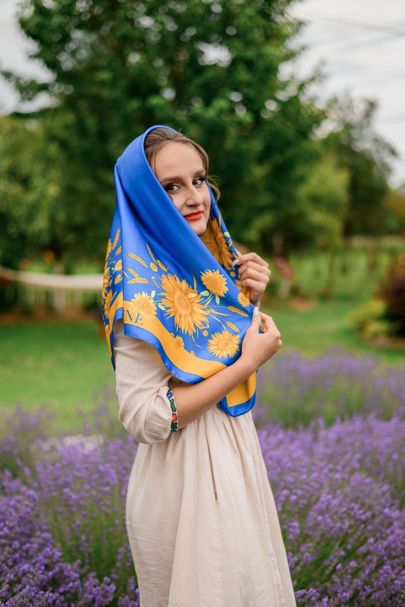 May include: A woman wearing a blue and yellow patterned scarf with sunflowers. She is standing in a field of lavender flowers.
