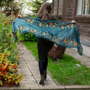 May include: A teal scarf with an orange floral pattern is held up in the air. The scarf is rectangular and appears to be made of a lightweight fabric. The floral design is detailed and adds a touch of elegance to the scarf. The scarf is being displayed outdoors.