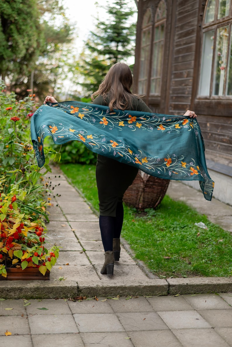 May include: A teal scarf with an orange floral pattern is held up in the air. The scarf is rectangular and appears to be made of a lightweight fabric. The floral design is detailed and adds a touch of elegance to the scarf. The scarf is being displayed outdoors.