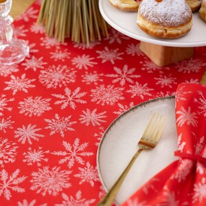 May include: A festive red table runner with a white snowflake pattern. A white plate with a gold fork and a matching napkin are set on the runner. A cake stand with pastries is in the background, suggesting a holiday theme.