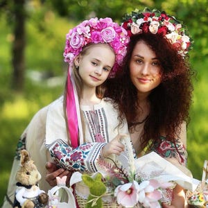 May include: A mother and daughter wearing floral crowns and traditional embroidered shirts. They are holding baskets filled with Easter decorations, including bunny figurines and flowers.