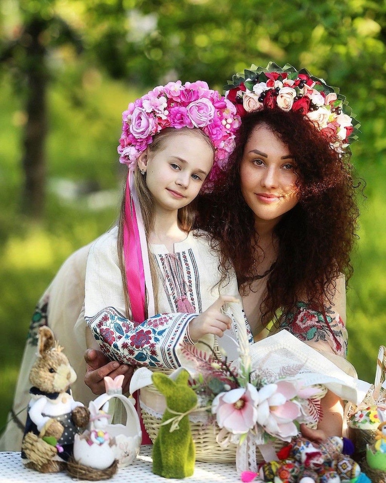 May include: A mother and daughter wearing floral crowns and traditional embroidered shirts. They are holding baskets filled with Easter decorations, including bunny figurines and flowers.