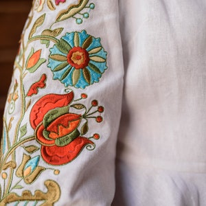 Op de afbeelding: Wit linnen stof met kleurrijke geborduurde bloemen in tinten rood, oranje, groen, blauw en geel.