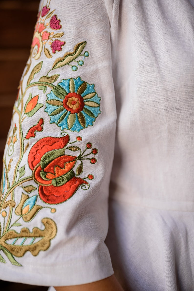 Op de afbeelding: Wit linnen stof met kleurrijke geborduurde bloemen in tinten rood, oranje, groen, blauw en geel.