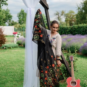 May include: A black scarf with an embroidered floral design featuring orange and green accents. The scarf is held up in front of a woman wearing a cream-colored dress with floral embroidery. A red acoustic guitar leans against a wooden structure.