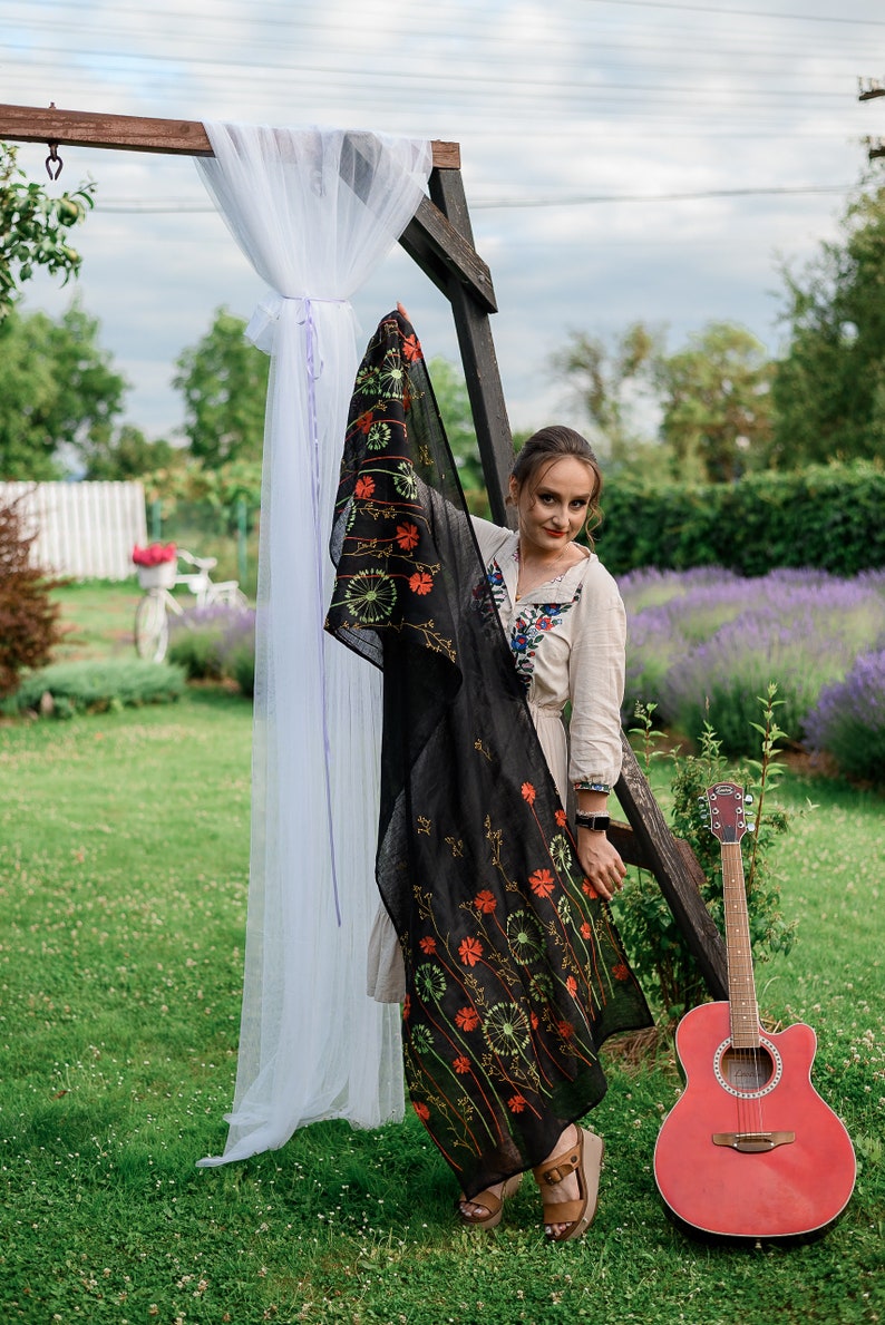 May include: A black scarf with an embroidered floral design featuring orange and green accents. The scarf is held up in front of a woman wearing a cream-colored dress with floral embroidery. A red acoustic guitar leans against a wooden structure.