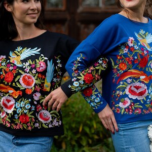 Puede incluir: Dos camisas de manga larga con bordados florales coloridos. Una camisa es negra con un dise&ntilde;o de p&aacute;jaro azul, y la otra es azul con un dise&ntilde;o de p&aacute;jaro rojo.