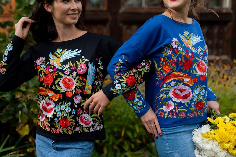 Puede incluir: Dos camisas de manga larga con bordados florales coloridos. Una camisa es negra con un dise&ntilde;o de p&aacute;jaro azul, y la otra es azul con un dise&ntilde;o de p&aacute;jaro rojo.