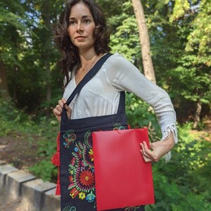 May include: A black tote bag with a colorful floral embroidery design and red tassels. The bag is being held by a person and has a red folder inside.