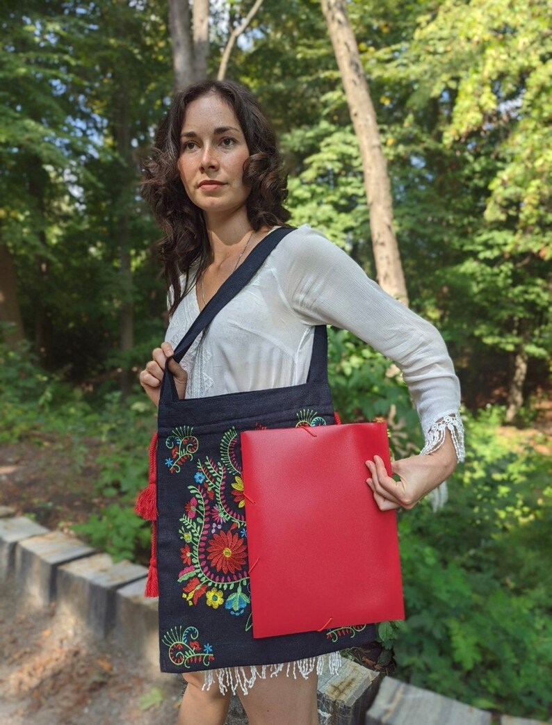 May include: A black tote bag with a colorful floral embroidery design and red tassels. The bag is being held by a person and has a red folder inside.