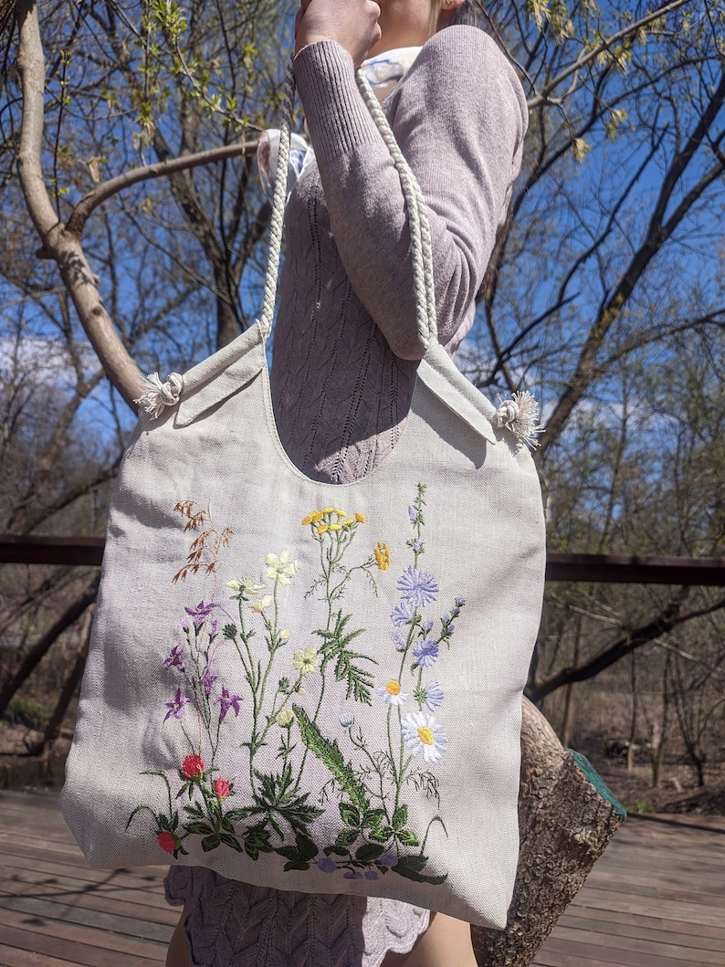 Op de afbeelding: Een witte canvas tote bag met geborduurde wilde bloemen in tinten van paars, geel, wit en groen. De tas heeft witte touwhandvatten.