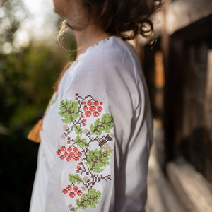 May include: A white embroidered blouse with a floral pattern in shades of green, red, and brown. The blouse has a gathered neckline and long sleeves.