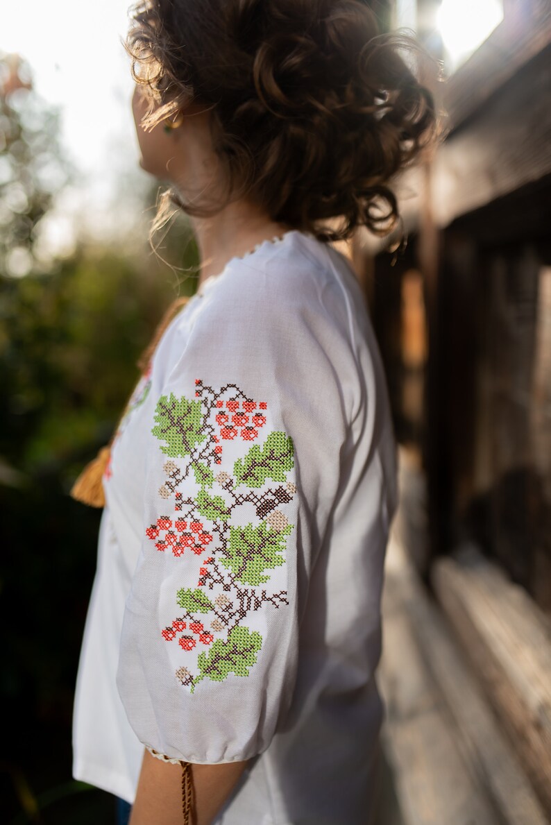 May include: A white embroidered blouse with a floral pattern in shades of green, red, and brown. The blouse has a gathered neckline and long sleeves.