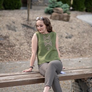 May include: A woman wearing a green sleeveless top with embroidered details sits on a wooden bench. She is wearing brown and white checked trousers and white trainers.