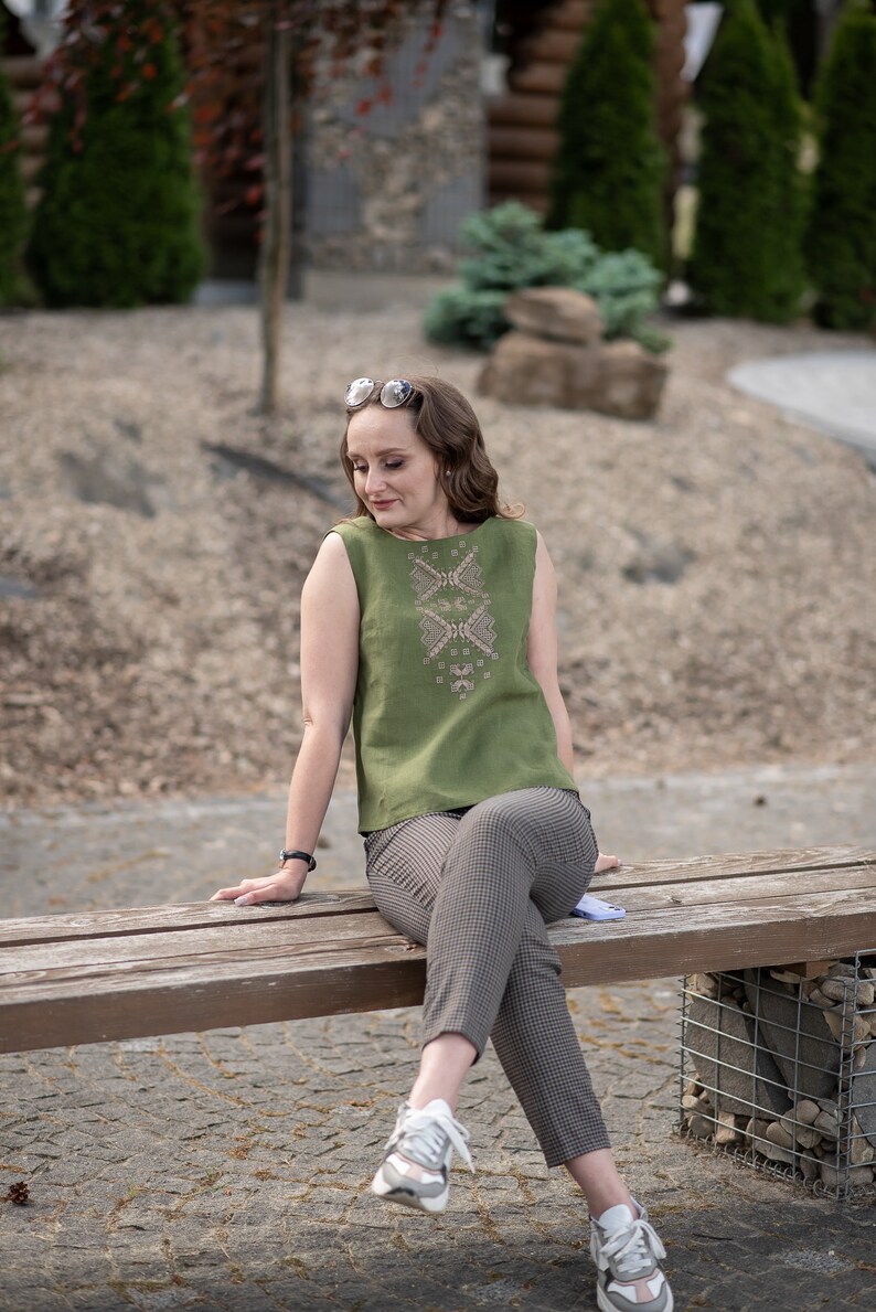 May include: A woman wearing a green sleeveless top with embroidered details sits on a wooden bench. She is wearing brown and white checked trousers and white trainers.