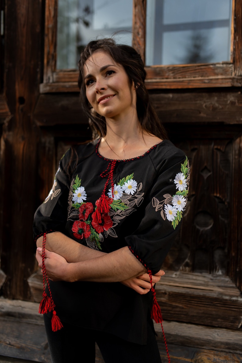 May include: A woman wearing a black embroidered blouse with red and white floral designs. The blouse has red tassels on the sleeves and neckline.