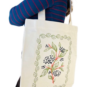 May include: A white canvas tote bag with a floral design embroidered on the front. The design features a red stem with green leaves and black berries. The design is framed by a green leafy border.