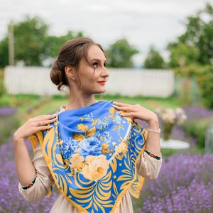 May include: A woman wearing a blue and yellow floral patterned scarf. The scarf has a yellow border and is tied around her neck. She is standing in a field of purple flowers.