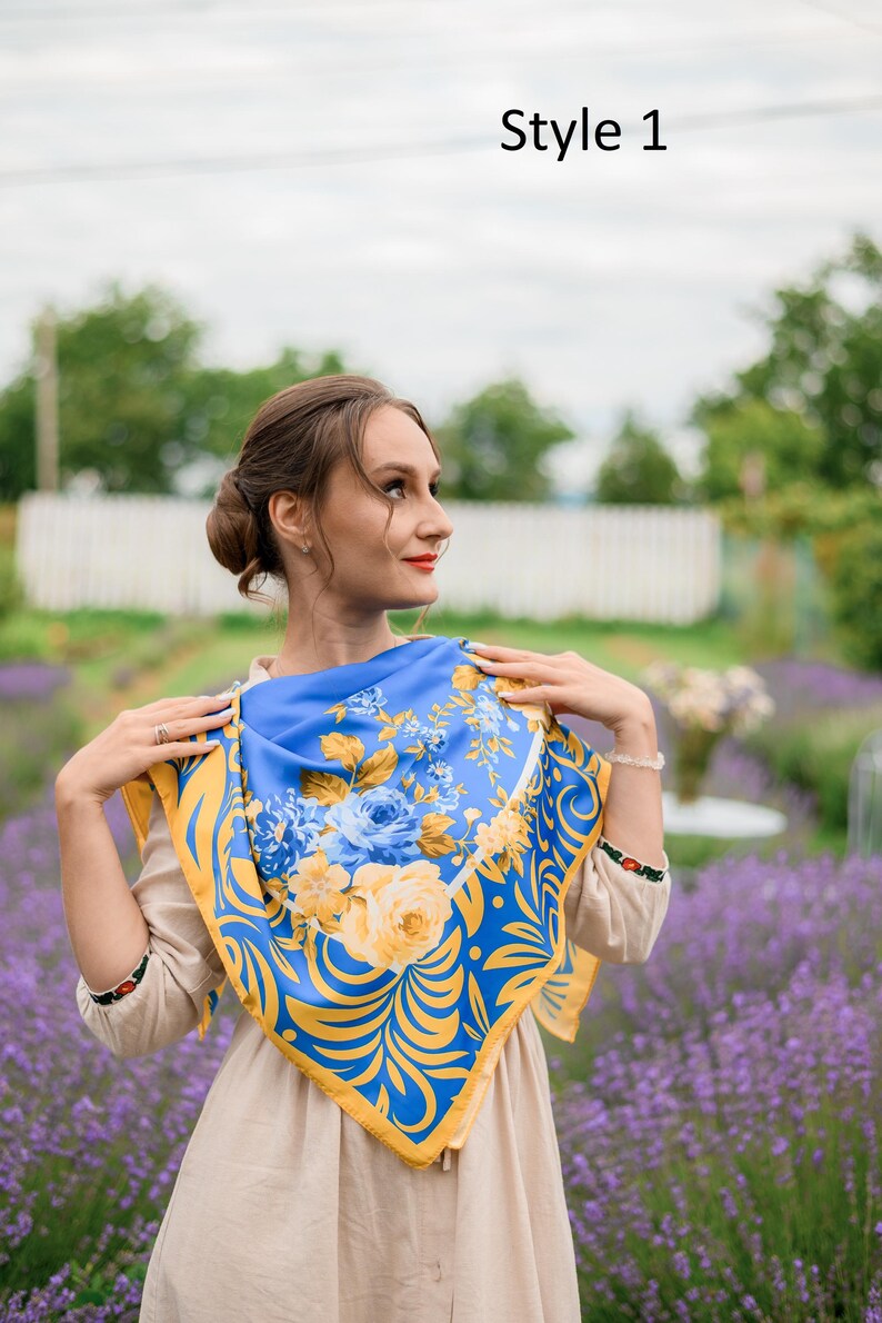 May include: A woman wearing a blue and yellow floral patterned scarf. The scarf has a yellow border and is tied around her neck. She is standing in a field of purple flowers.