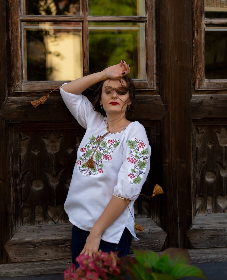 May include: A woman with long brown hair wearing a white embroidered blouse with red and green floral designs and blue jeans. She is standing in front of a wooden door with a window.