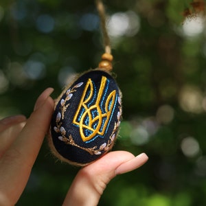 K&ouml;nnte beinhalten: Ein blaues und golden besticktes Ei mit einem ukrainischen Dreizack-Symbol. Das Ei h&auml;ngt an einer Schnur und wird von einer Hand gehalten.