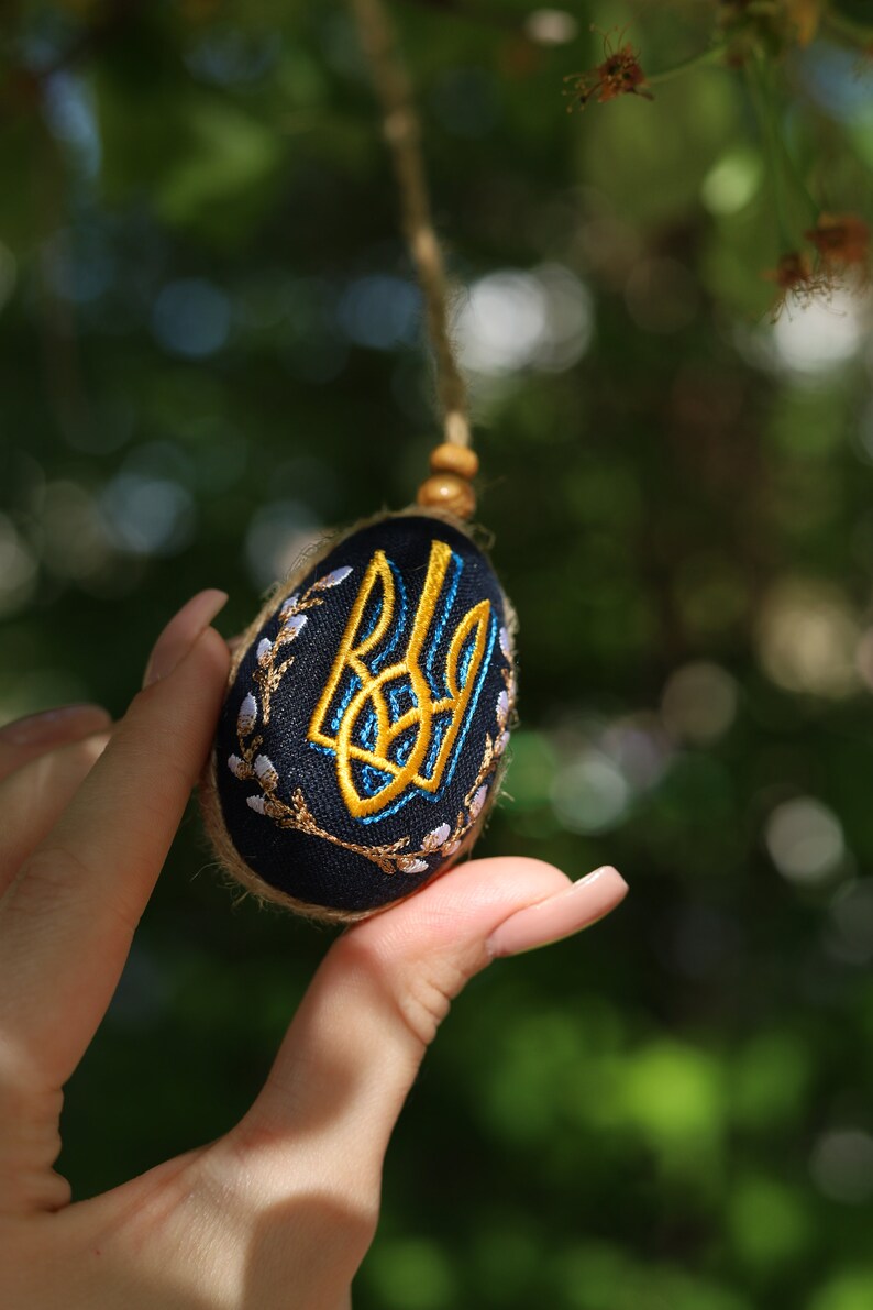 K&ouml;nnte beinhalten: Ein blaues und golden besticktes Ei mit einem ukrainischen Dreizack-Symbol. Das Ei h&auml;ngt an einer Schnur und wird von einer Hand gehalten.