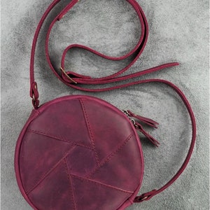 May include: A round, burgundy leather crossbody bag with a geometric design on the front. The bag features a long, adjustable strap and a zipper closure. The bag's diameter is approximately 8 inches.
