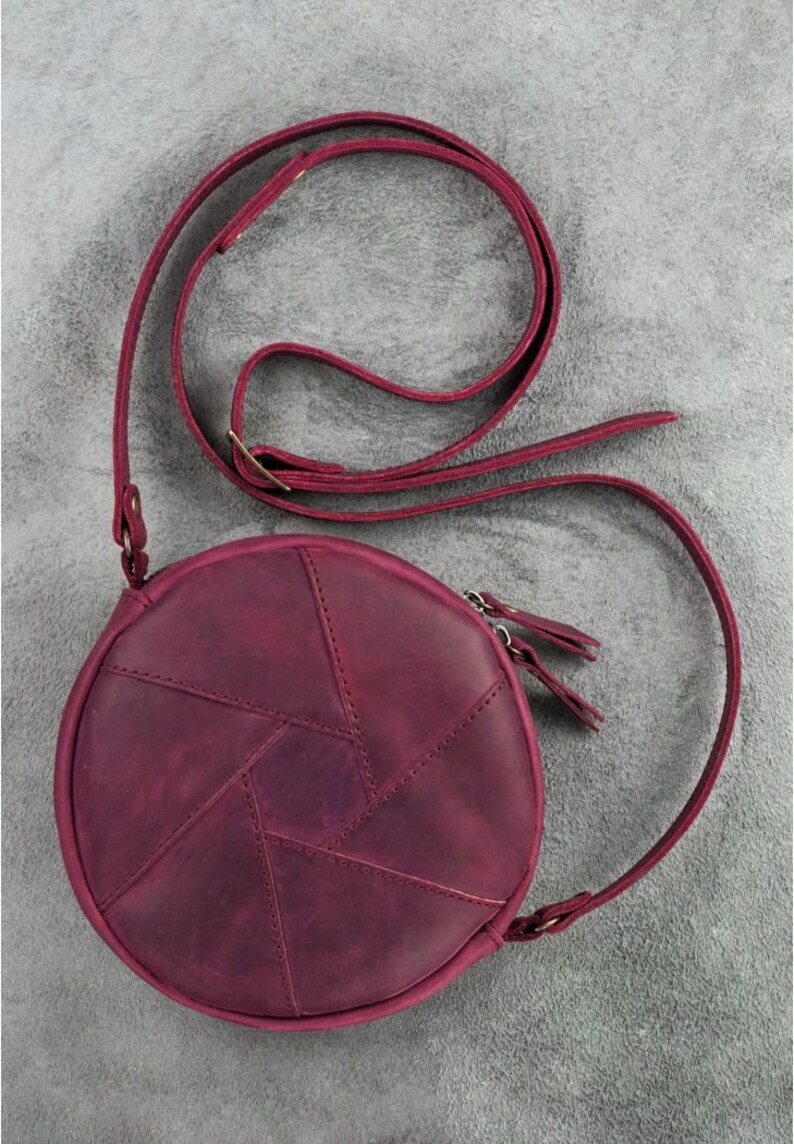 May include: A round, burgundy leather crossbody bag with a geometric design on the front. The bag features a long, adjustable strap and a zipper closure. The bag's diameter is approximately 8 inches.