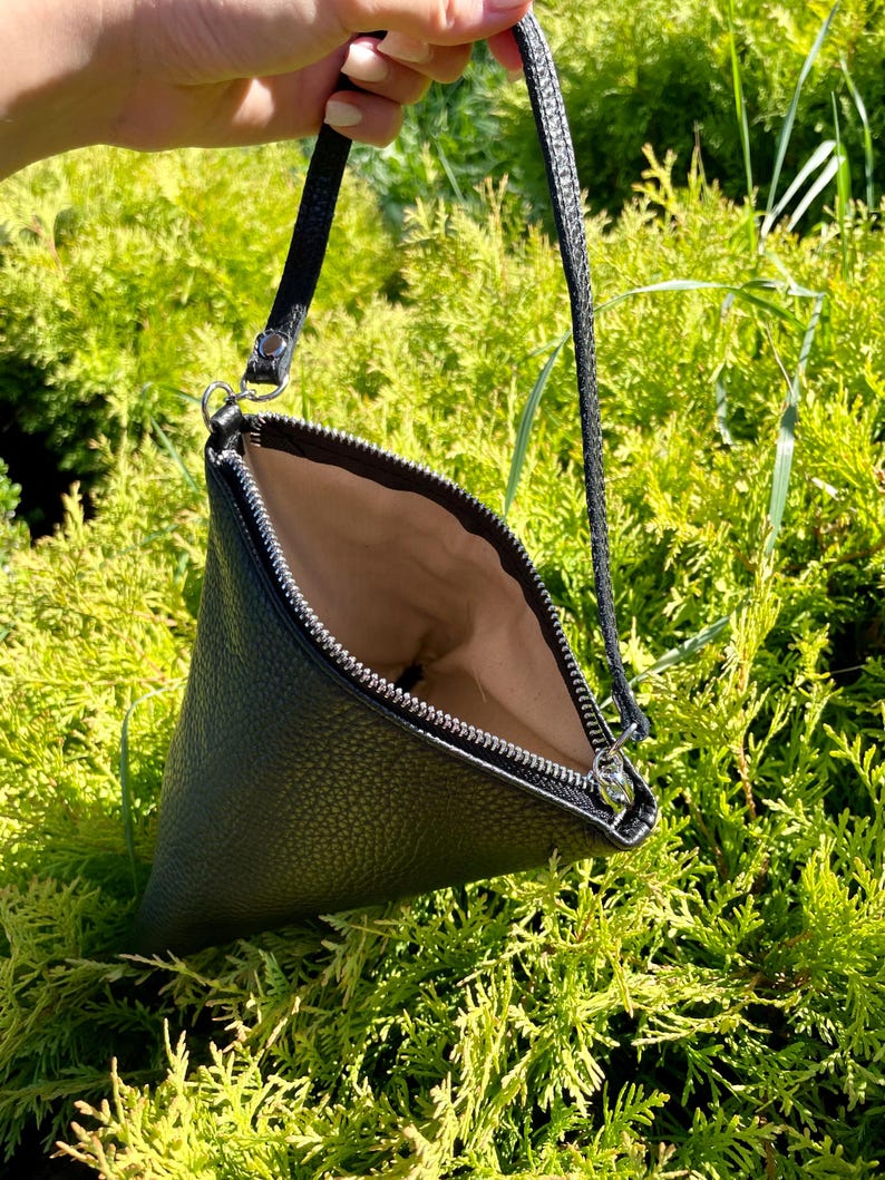 May include: A black leather triangular shaped clutch bag with a zipper closure and a long strap.