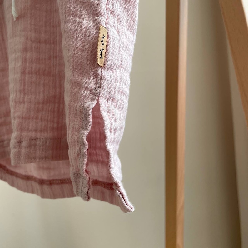 May include: A pink cotton gauze top with a small brown tag that says "xxx" on it. The top has a side slit and a hemmed edge.