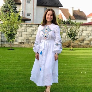 Może przedstawiać: Biała sukienka lniana z haftowanymi geometrycznymi wzorami w kolorze niebieskim, pomarańczowym i zielonym. Sukienka ma długie rękawy, dopasowany gorset i rozkloszowaną sp&oacute;dnicę.