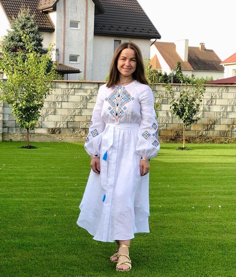 Może przedstawiać: Biała sukienka lniana z haftowanymi geometrycznymi wzorami w kolorze niebieskim, pomarańczowym i zielonym. Sukienka ma długie rękawy, dopasowany gorset i rozkloszowaną sp&oacute;dnicę.