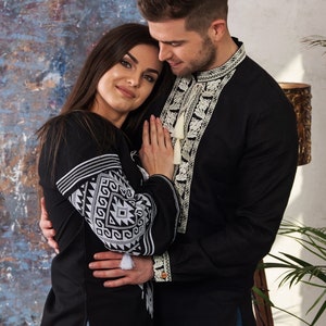 May include: A couple wearing black shirts with white embroidered details. The shirts have intricate patterns on the sleeves and around the neckline. The woman is wearing blue jeans. The man's shirt has a tassel.