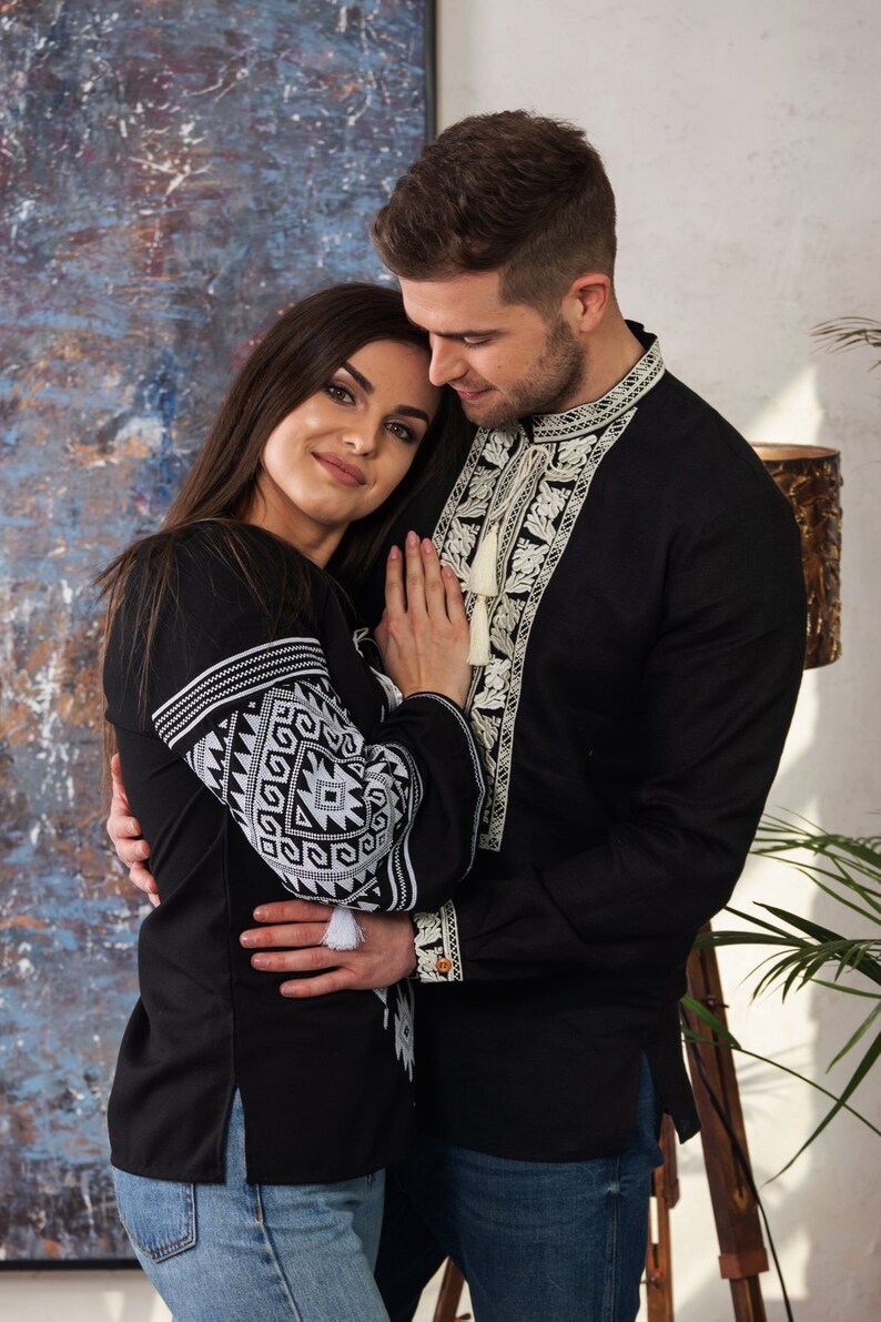 May include: A couple wearing black shirts with white embroidered details. The shirts have intricate patterns on the sleeves and around the neckline. The woman is wearing blue jeans. The man's shirt has a tassel.
