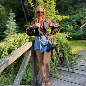 May include: A woman wearing a black embroidered blouse with red and white accents, a denim skirt, and brown sandals. She is standing on a wooden bridge with green foliage in the background.