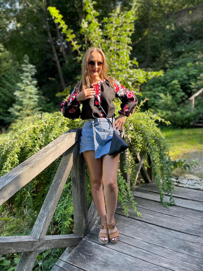 May include: A woman wearing a black embroidered blouse with red and white accents, a denim skirt, and brown sandals. She is standing on a wooden bridge with green foliage in the background.