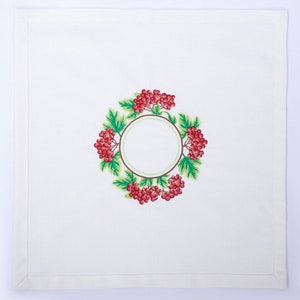 May include: White linen napkin with a detailed embroidered design. The design features a circular wreath of red berries and green leaves surrounding a blank circle, perfect for personalization.