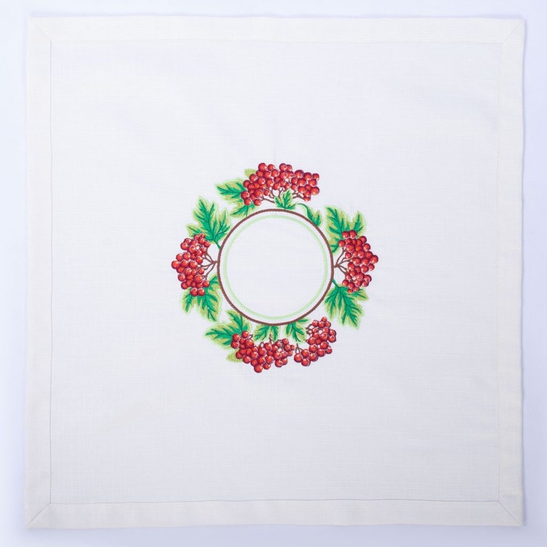 May include: White linen napkin with a detailed embroidered design. The design features a circular wreath of red berries and green leaves surrounding a blank circle, perfect for personalization.