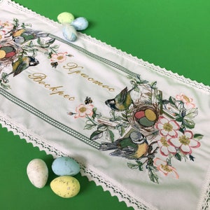 以下が含まれることがあります： 白い刺繍テーブルランナー、花と鳥のデザイン、テキスト「Христос Воскрес」が特徴です。 ランナーはレースのトリムが付いており、イースターに最適なカラフルな卵で飾られています。