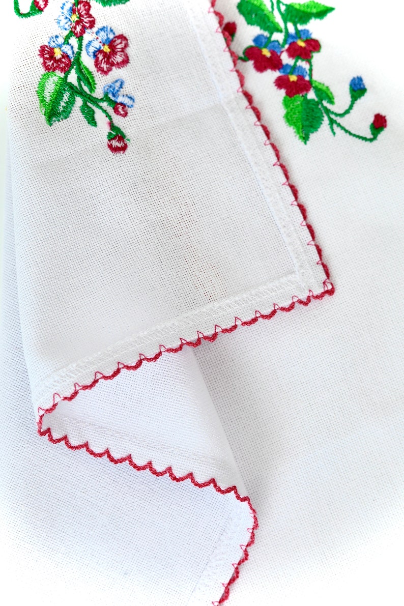 Pode incluir: Tecido branco com uma borda bordada vermelha e um desenho floral em vermelho, verde e azul.