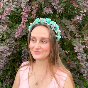 May include: A woman wearing a floral crown with light blue and silver accents. The crown is made of fabric flowers and is set against a backdrop of green and purple foliage. She is wearing a pink sleeveless top and a silver necklace with a yellow pendant.