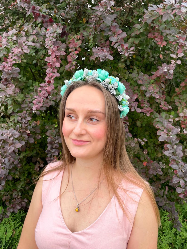 May include: A woman wearing a floral crown with light blue and silver accents. The crown is made of fabric flowers and is set against a backdrop of green and purple foliage. She is wearing a pink sleeveless top and a silver necklace with a yellow pendant.
