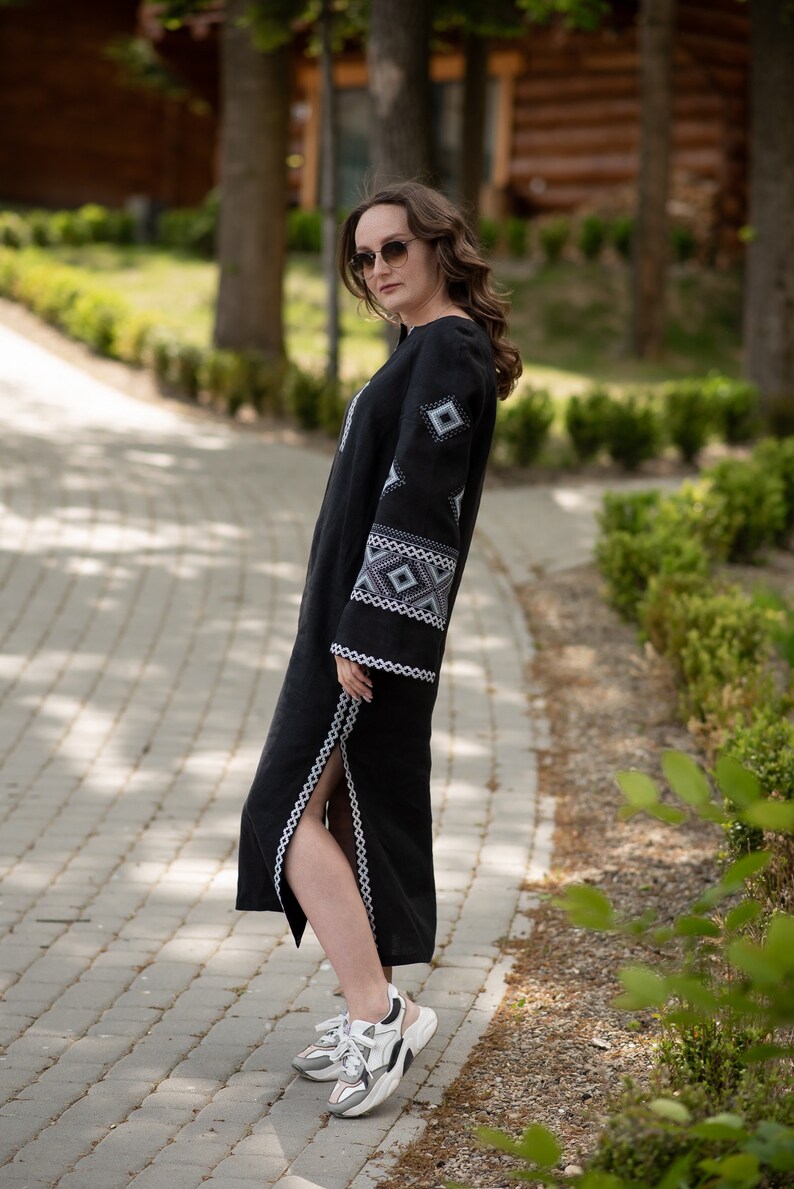 Pode incluir: Vestido longo de linho preto com padr&otilde;es geom&eacute;tricos bordados em branco e cinza nas mangas e no corpete. O vestido tem uma fenda alta e &eacute; usado com t&ecirc;nis brancos e cinza. O vestido &eacute; longo maxi.