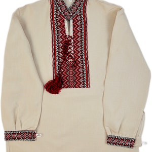 Op de afbeelding: Een beige shirt met lange mouwen en ingewikkelde rode en zwarte borduursels op de kraag, borst en manchetten. Het shirt heeft een decoratieve kwast en een veterhalslijn, met een traditioneel volksontwerp.