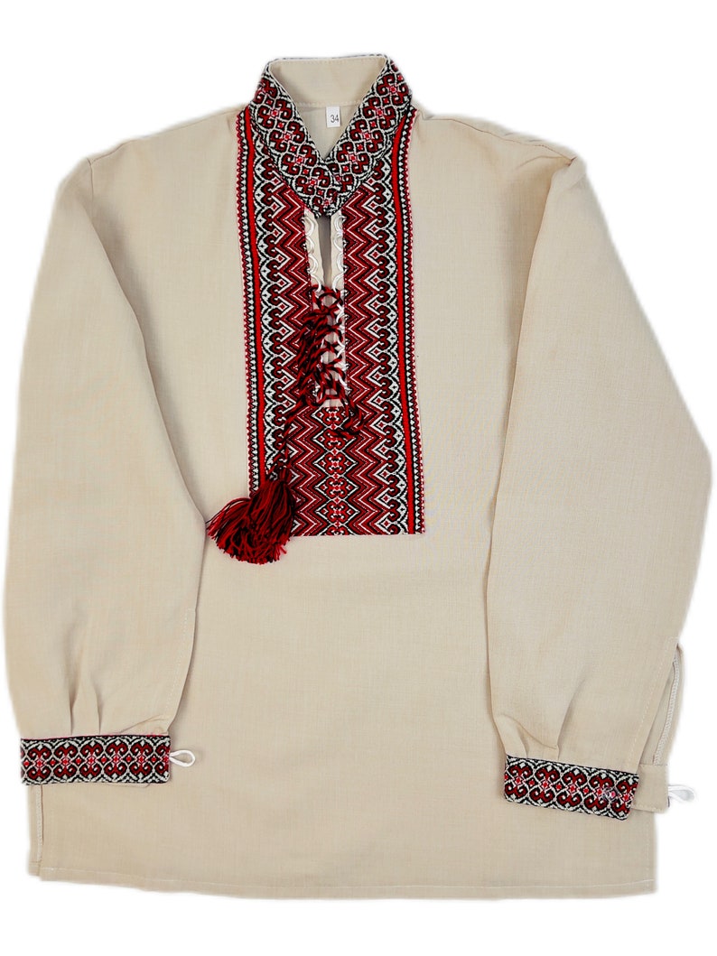 Op de afbeelding: Een beige shirt met lange mouwen en ingewikkelde rode en zwarte borduursels op de kraag, borst en manchetten. Het shirt heeft een decoratieve kwast en een veterhalslijn, met een traditioneel volksontwerp.